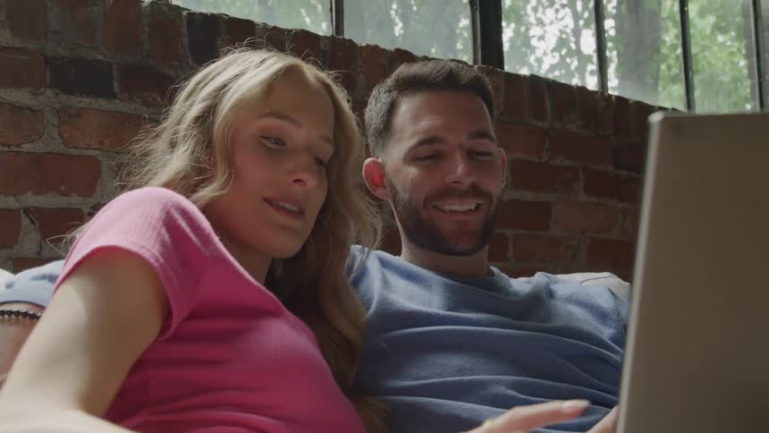 Couple cuddled on the couch smiling while looking at a laptop