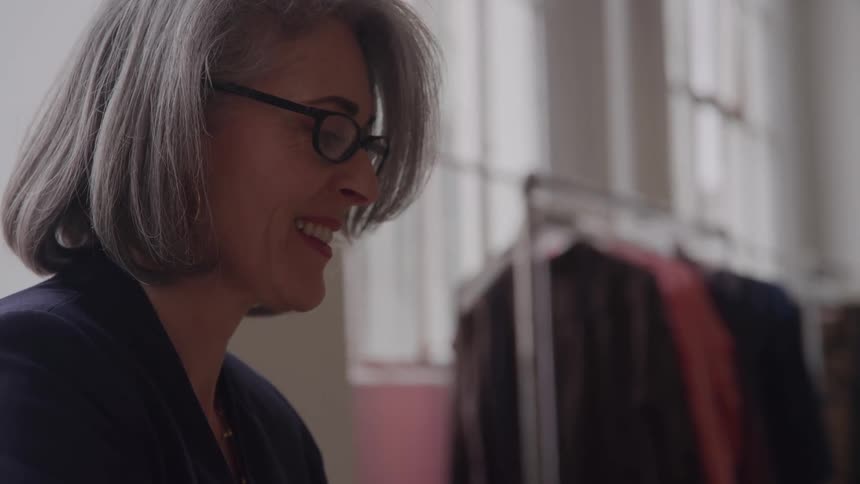 A grayhaired woman wearing glasses smiles warmly in a boutique with clothing racks behind her