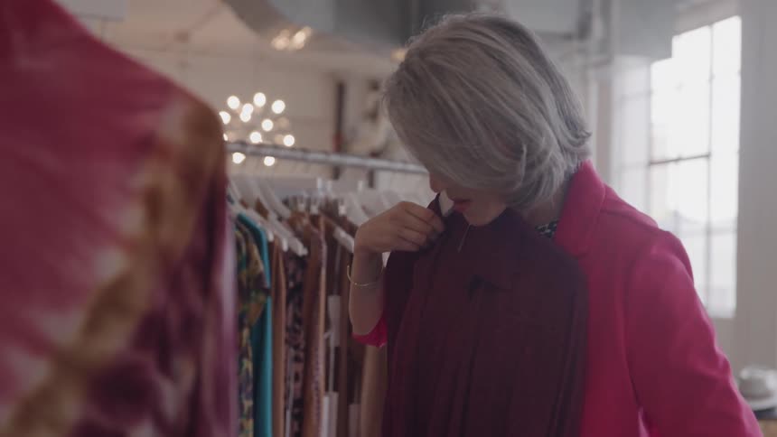 The video clip features an older woman with short gray hair and a bright pink blazer browsing clothes on a rack in a boutique she pauses to examine a dark red blouse closely before turning away from the camera