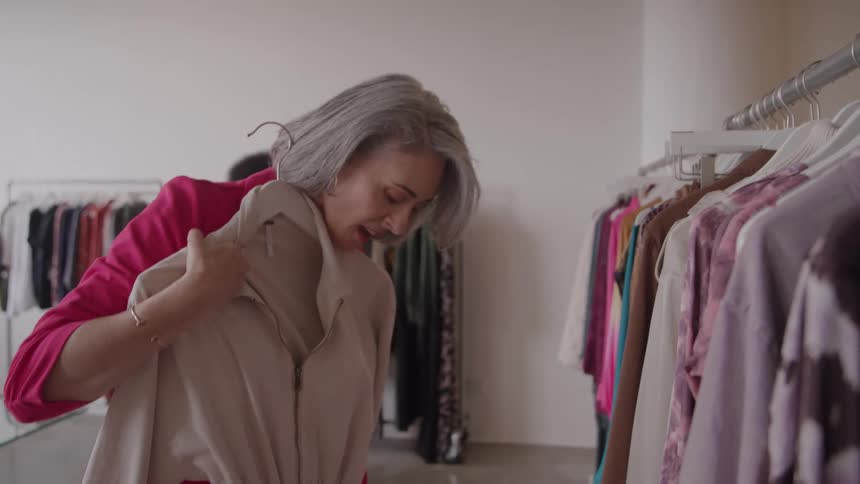 A woman in a pink jacket reaches up to a clothing rack and removes a light brown garment on a hanger then holds it up to her face and examines it with a smile in a clothing store