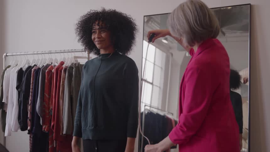 A young woman stands in front of a mirror inside a clothing shop as another woman uses a measuring tape to take her measurements focusing carefully to ensure a precise fit