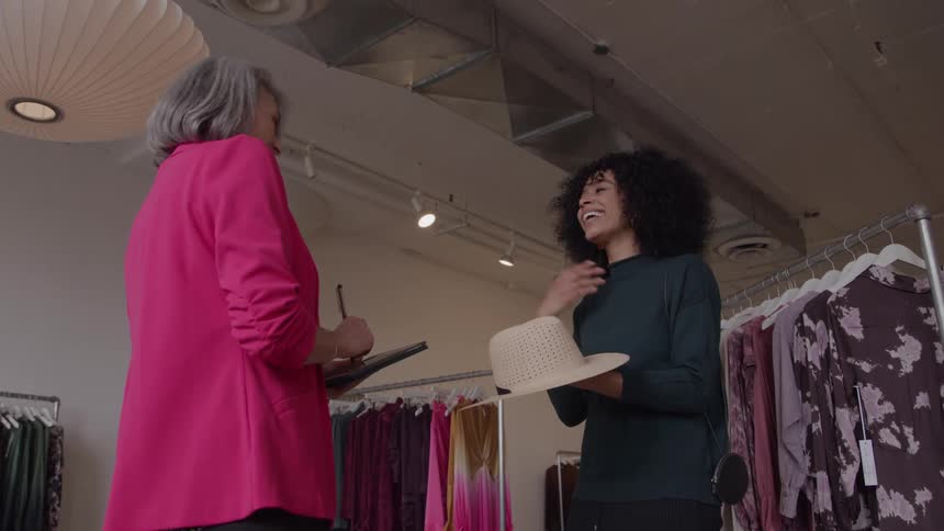 The video clip features two women in a clothing store engaged in a conversationan older woman in a bright pink blazer is talking to a younger woman with curly hair who is holding a straw hat