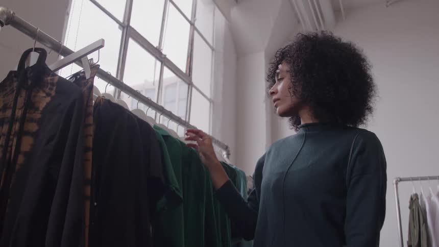 A woman with curly dark hair stands next to a metal clothing rack and slides her hand along the hanging garments while facing a large multipane window in an indoor retail space