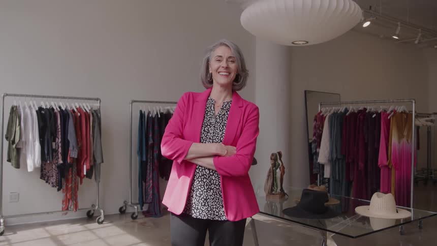 A woman with gray hair and a pink blazer stands with arms folded in the center of a retail store with clothing racks and a display table on either side smiling directly toward the camera