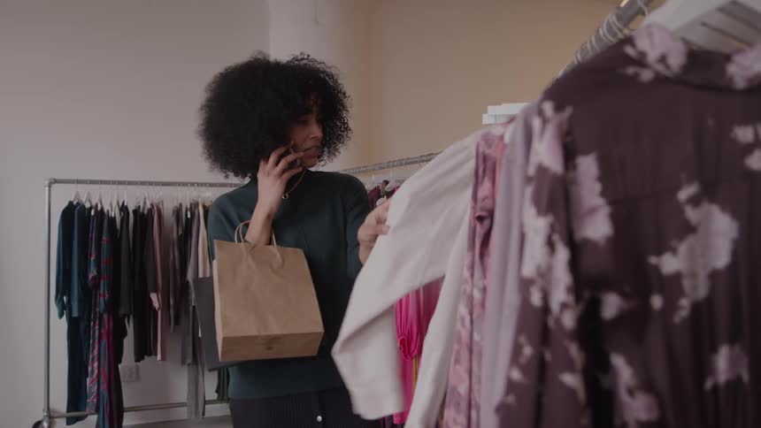 The woman is shopping in the clothing store as she talks on her phone