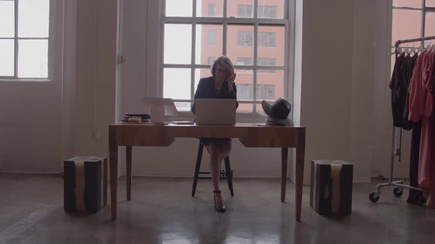 Grayhaired woman working on a laptop at a wooden desk inside a bright fashion studio