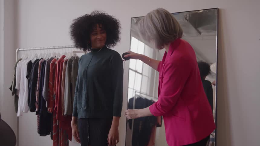 The video shows an older woman in a pink blazer helping a younger woman with curly hair try on a garment in a clothing store the older woman who appears to be a store employee is smoothing the fabric on the customers shoulder near a large mirr