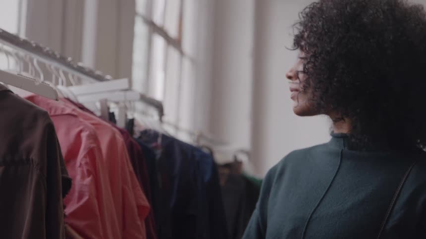 The video features a young woman with curly hair browsing a rack of clothes in a boutiqueshe runs her hand over the garments taking a moment to look closely at a light pink top or dress