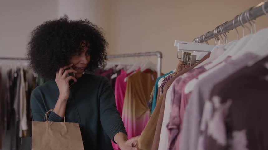 A curlyhaired woman smiles while talking on the phone holding a shopping bag as she browses a rack and fingers a tan garment