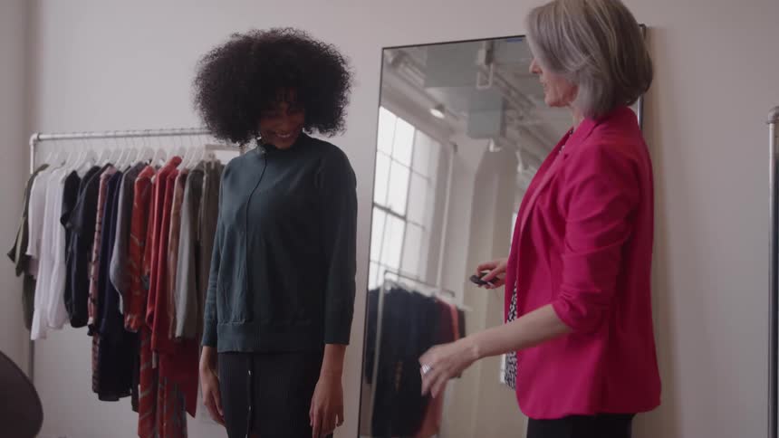 A smiling woman gets her sleeve measured by a grayhaired stylist in a boutique with a floor mirror and clothing racks behind them