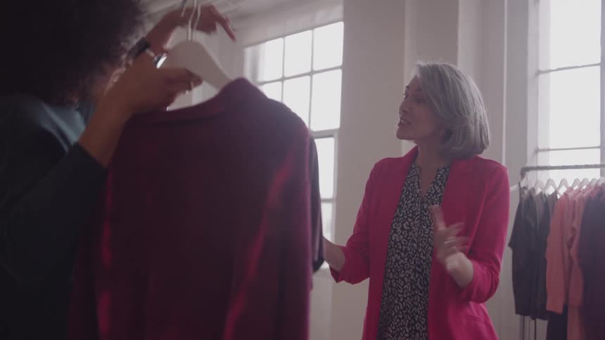 A woman laughs while holding a burgundy sweater to her neck as a grayhaired stylist in a pink blazer smiles beside her