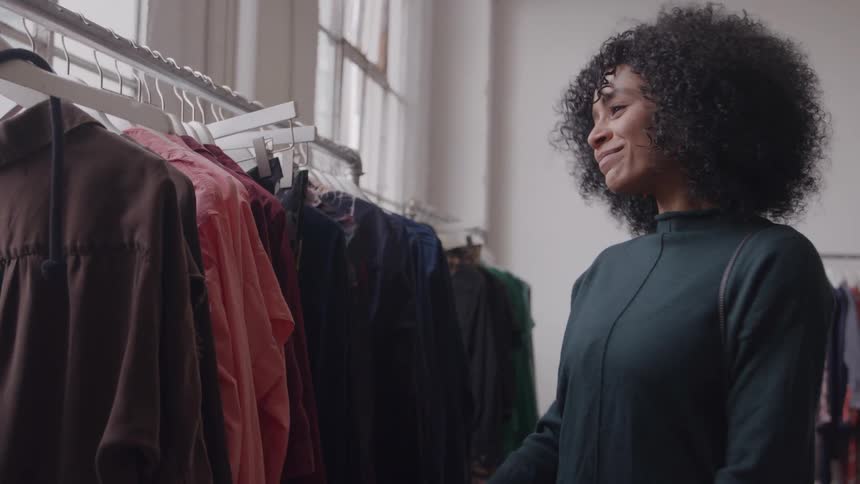 Woman with curly hair browses a clothing rack gently touching garments with a calm pleased expression