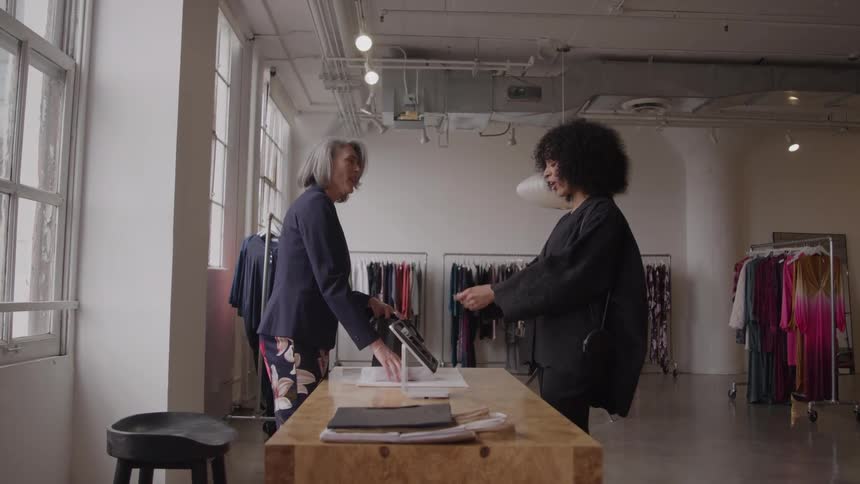 Grayhaired shop sales associate smiles as she hands a shopping bag to a curlyhaired customer across the checkout table in a bright boutique