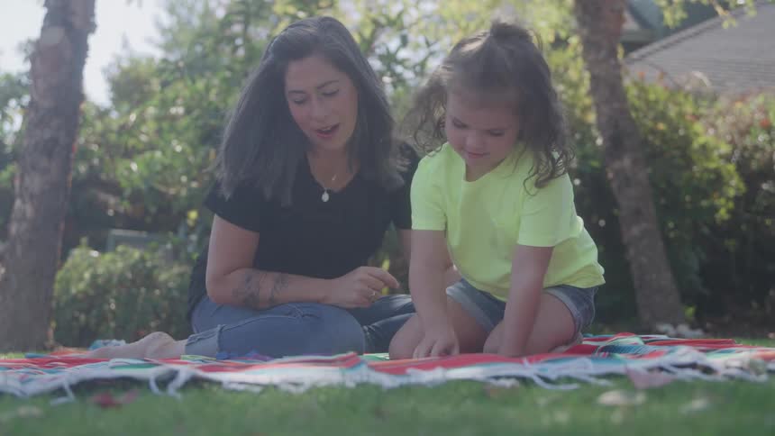 Woman sitting on the grass with a child pointing at a game on the blanket