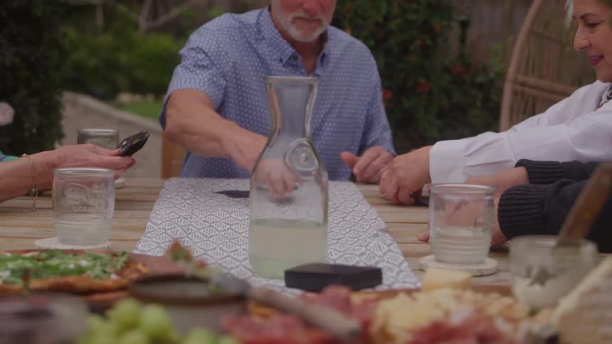 The video clip is a closeup shot of four peoples hands around a wooden outdoor dining table suggesting a social meal the table is set with food like pizza and charcuterie a glass pitcher of lemonade and individuals are holding or reaching for glasse