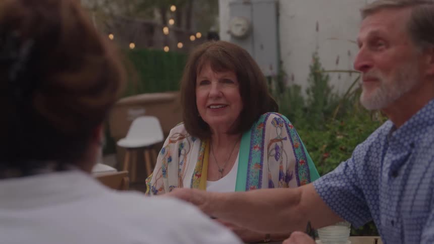 A middleaged woman in a colorful printed shawl and a graybearded man in a blue collared shirt sit outdoors at a table talking and playing cards with an unseen third person with string lights and a green garden visible in the blurred background