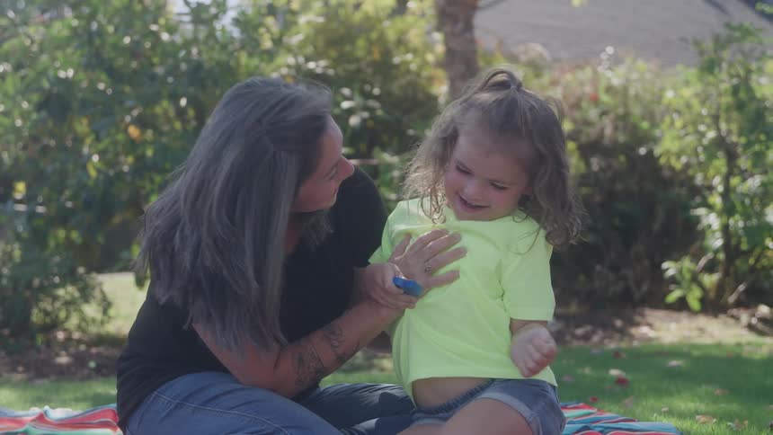 An adult woman with long gray hair sits on a picnic blanket on the grass engaging in playful interaction with a young child who is laughing while touching the childs chest in a sunny outdoor park
