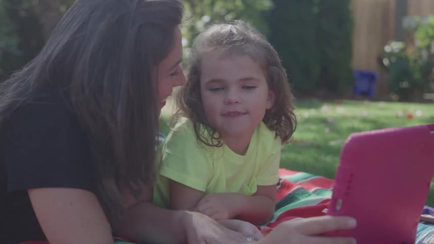 Woman and child lying on a blanket look at a pink tablet