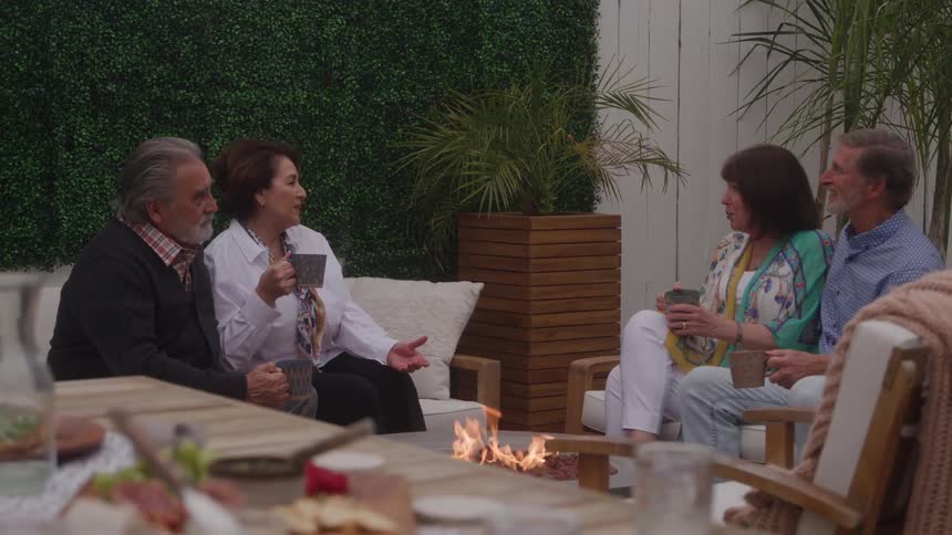 Four adults sit on outdoor lounge chairs and a couch around a small fire pit all holding mugs and engaging in conversation and laughter while a wooden dining table with food in the foreground