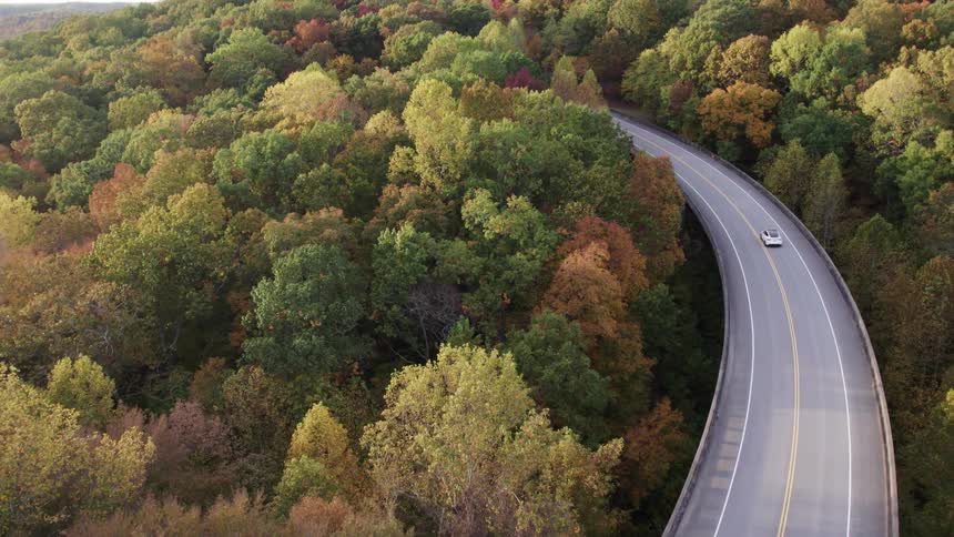 An overhead shot captures a curving elevated roadway with a white car passing by running through a hilly landscape covered in dense forest trees