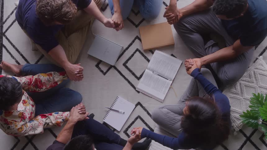 An overhead shot shows six people sitting on a patterned rug in a circle with hands joined with books and notebooks in the center as the group appears to be praying