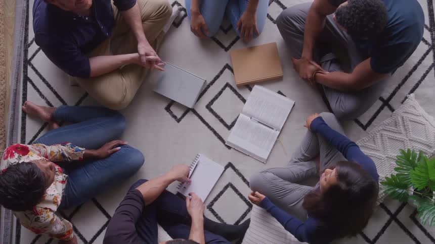 An overhead shot shows six people sitting on the floor in a circular formation on a patterned rug with open and closed books and a notebook resting in the center as the group appears to be engaged in a discussion