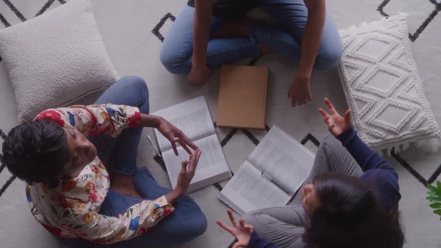 An overhead shot shows three people sitting on the floor in a circular arrangement with open books in front of them with one woman making hand gestures as she talks during a group discussion