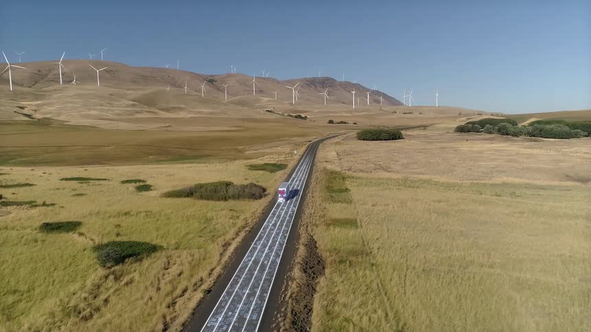 Red truck drives along a road surrounded by fields with wind turbines in the background