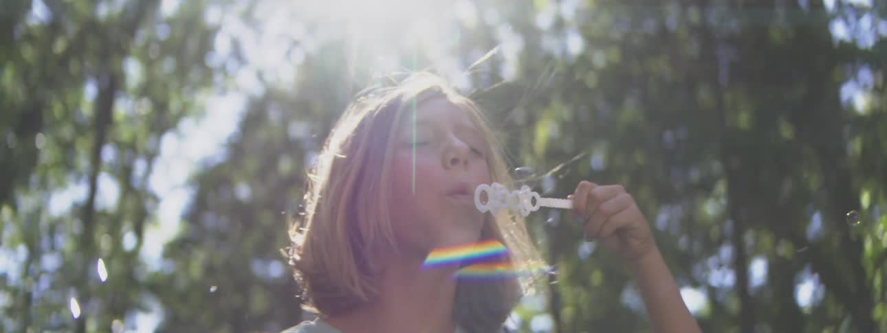 Person blowing soap bubbles under sunlight among trees