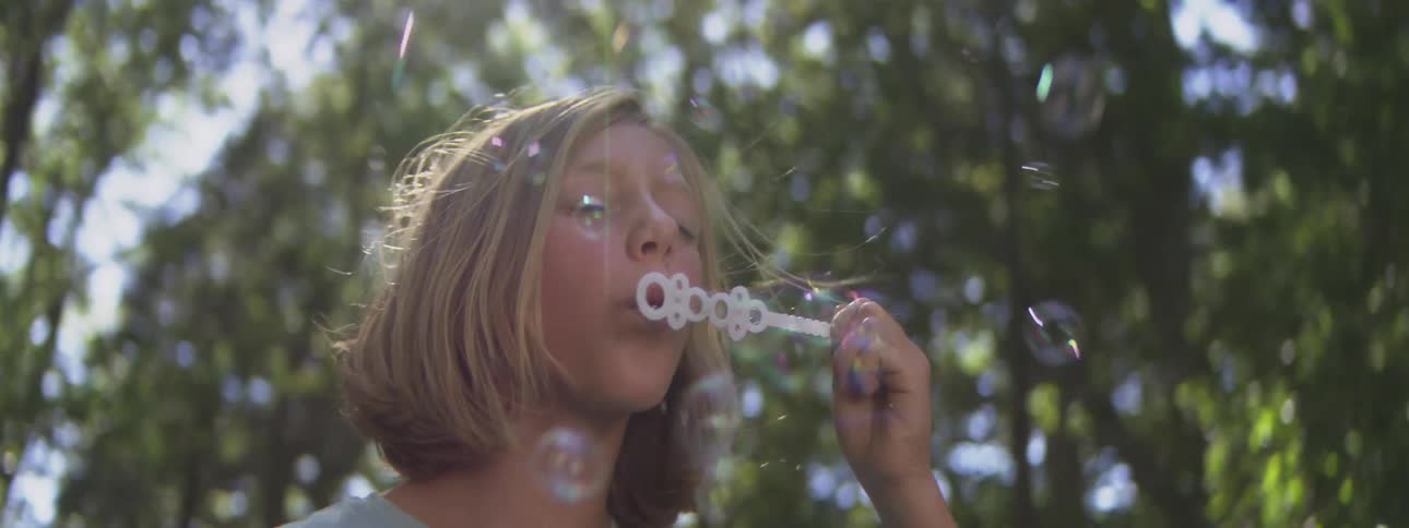 A blonde boy holds bubble wand apparatus to mouth and exhales creating multiple soap spheres that float into the air while standing among tall plants