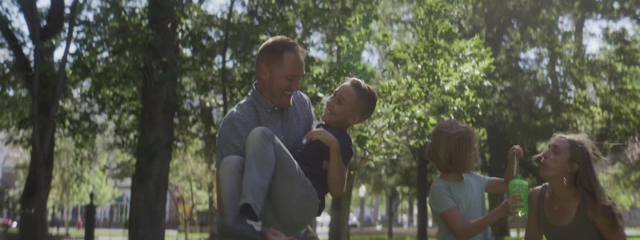 Four people stand on grass in a treed area the man lifts the boy off the ground and then returns him to the ground while the girl blows soap spheres and the woman stands nearby all engaging in the activity