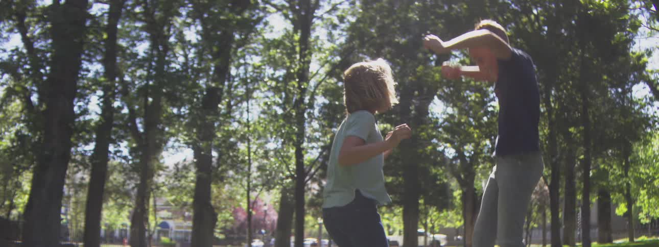 Two children a boy in dark clothes and a child in light clothes jump and move their arms and bodies rapidly while facing each other in a sunny outdoor setting with dense foliage in the background