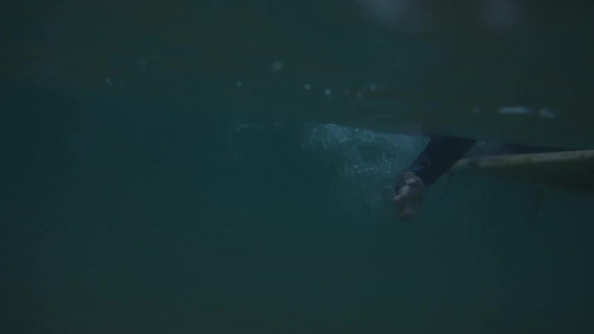 A camera is positioned beneath the water surface capturing the underside of a surfboard and the arm of a person wearing dark clothing moving through the water to propel the board forward