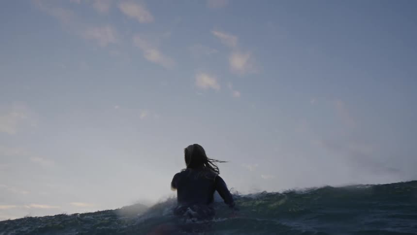 The video is an extreme lowangle shot of a person paddling a surfboard out over a wave at dusk or dawn the cloudy sky is a soft blend of pink gray and yellow providing a muted warm backlight to the silhouetted water and surfer