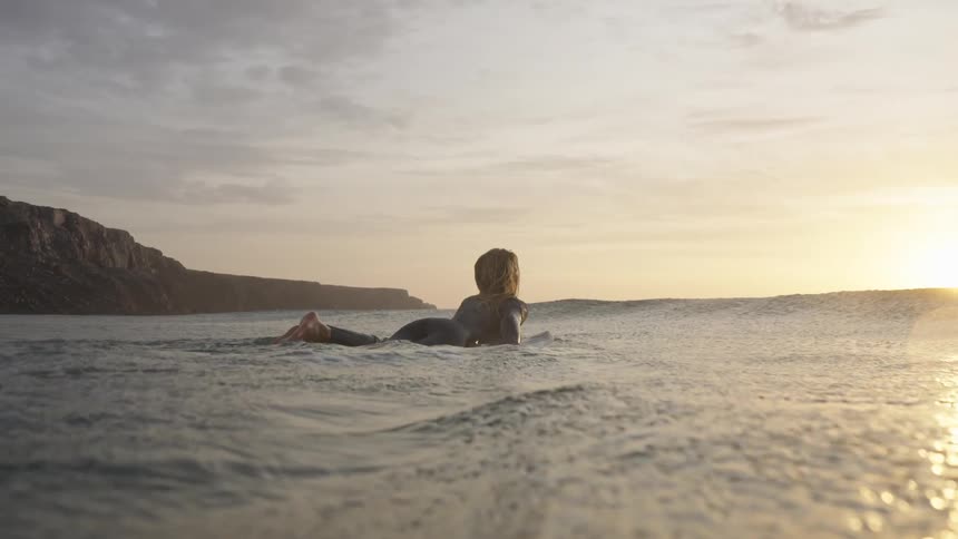 A woman in a wetsuit paddles on a surfboard toward golden light with rocky cliffs stretching along the shore