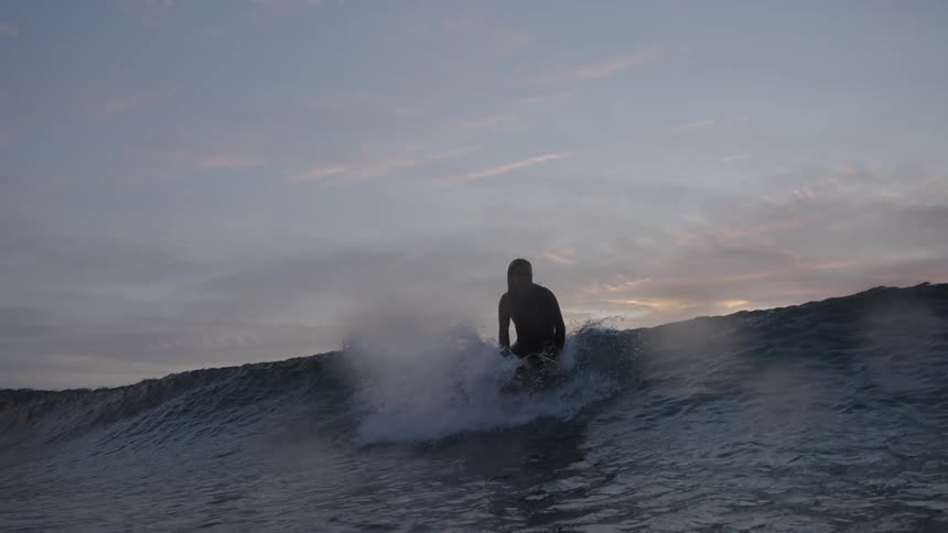 A surfer in a wetsuit rides a wave at the beach under a dramatic sky at dusk