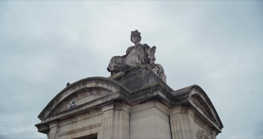 A weathered stone statue sits atop a grand neoclassical archway under a pale overcast sky