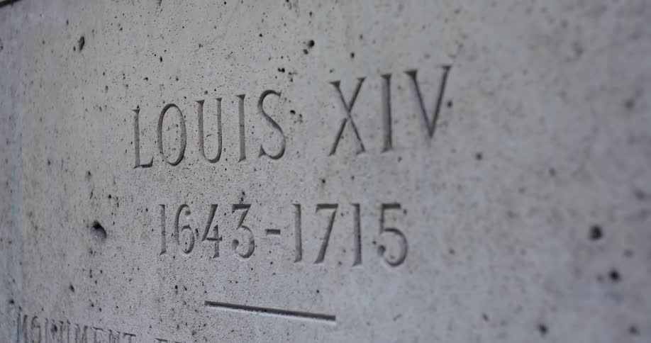 A camera is focused on the rough surface of a grey tombstone where the name louis xiv is carved in capital letters above the lifespan dates 16431715 which are followed by a short horizontal line