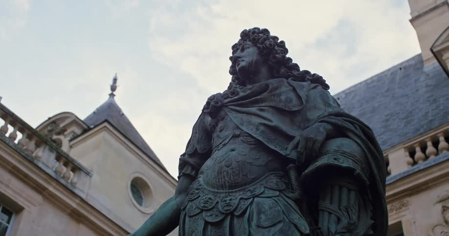 A bronze statue of a robed armored figure stands in a european courtyard with mansard roofs and a round window