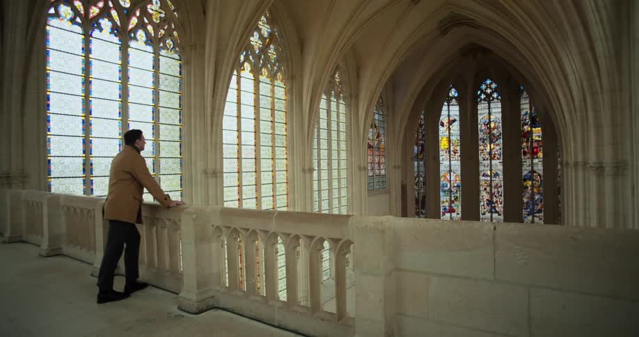 A man stands on an upper stone gallery within a gothic chapel looking at the towering ornate stained glass windows