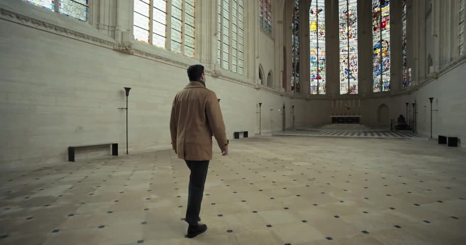 A man wearing a tan wool coat and dark pants walks slowly across the checkerboard tiled floor of a bright empty church or chapel looking up at the high walls lined with large arched stained glass windows and plain gothic windows