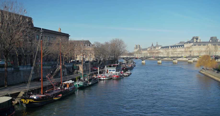 A river flowing from the foreground toward the background with multiple boats docked along the left bank beneath the quay wall and large lightcolored historical buildings under a clear sky