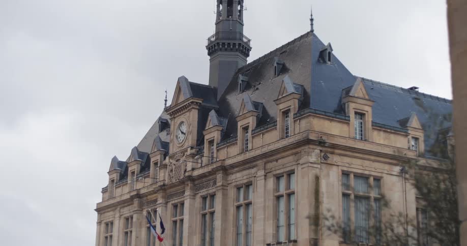 The video is an exterior shot of a historic building in paris with a prominent central spire and a clock face below it the architecture features a steep dark roof and lightcolored stone all set against a very bright overcast sky
