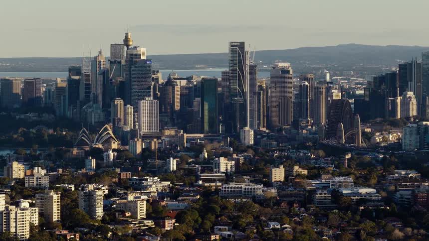 The video is a high wide shot of a dense modern city skyline with tall skyscrapers clustered together in the foreground there are rows of white residential buildings and trees below the commercial highrises possibly showing sydney australia