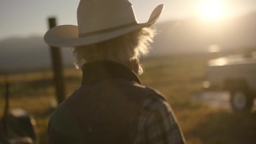 An old man is standing looking around on a ranch at sunset