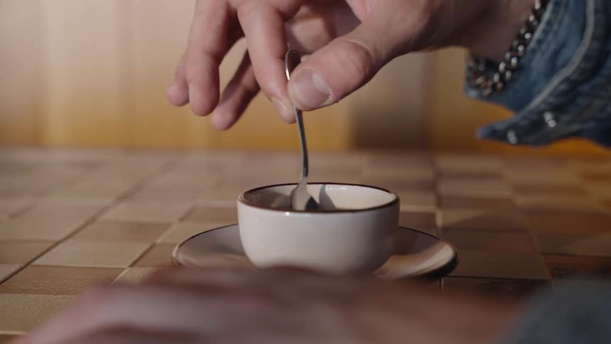 A persons hand holds a metal spoon and moves it inside a small white cup containing a dark liquid which rests on a saucer on a patterned surface