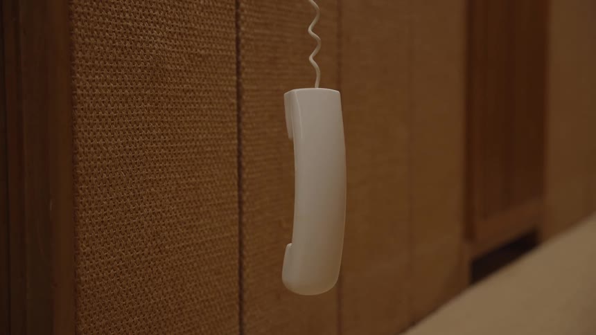 A white telephone receiver hangs suspended by its coiled cord against a woven textured wall panel