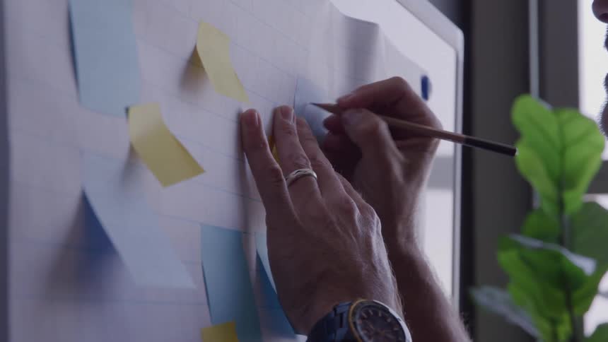 A persons hands hold a wooden pencil and draw lines on a white board decorated with multiple small colored adhesive papers while the person is wearing a wristwatch and a ring with a green indoor plant partially visible on the right