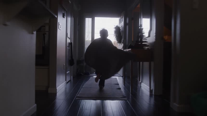 The video is an interior shot of a person carrying a long thick wooden plank through a dimly lit hallway the light wood slab contrasts with the dark flooring and walls of the room