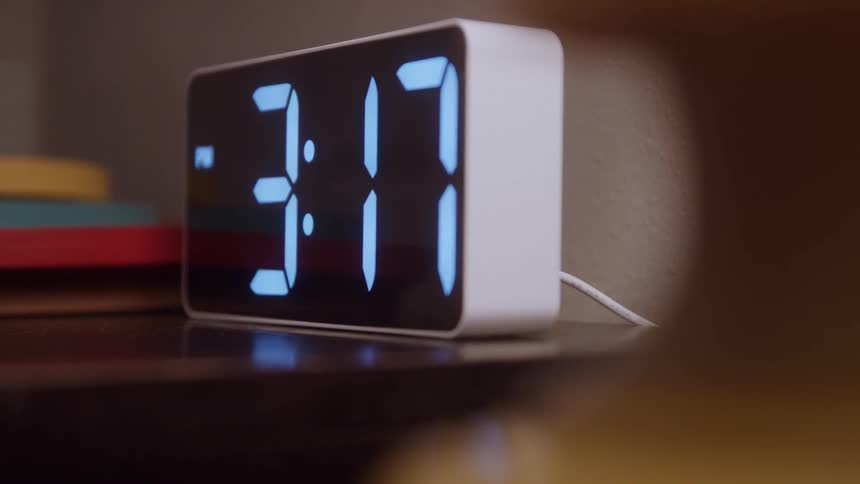A white digital alarm clock sits on a dark wooden nightstand displaying the time 317 in large bright blue numbers in a slightly out of focus shot suggesting an early morning view